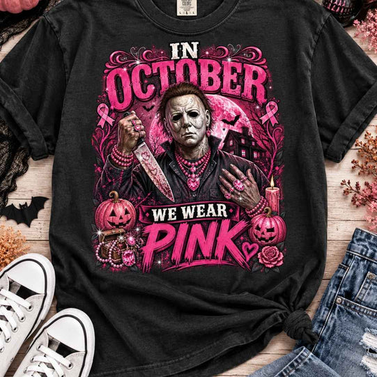 Black Michael Myers In October We Wear Pink Halloween graphic T-shirt with pink lettering, ribbons, pumpkins, and a haunted house.
