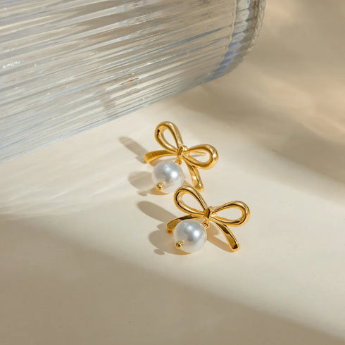 Gold-Tone Bow Earrings with Synthetic Pearls: Gold-tone bow earrings with white bead drops beside a glass vase